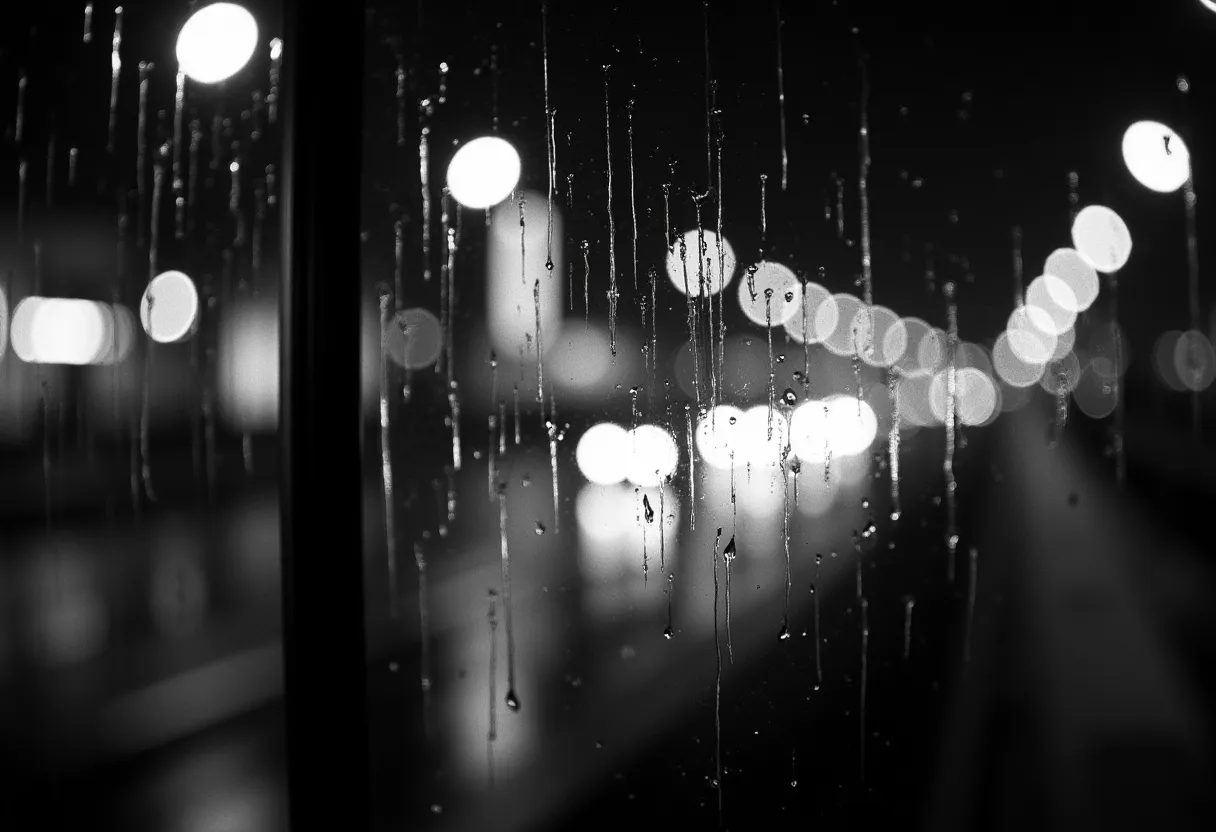 A monochrome night photograph taken through a rain-covered window. In the foreground the glass is in sharp focus, beaded with droplets and streaked by long vertical runnels of water, with a dark window frame at the left edge. Beyond the wet glass a city at night is thrown far out of focus, soft round bokeh from streetlights and lit windows, indistinct dark buildings and a railing receding to the right.